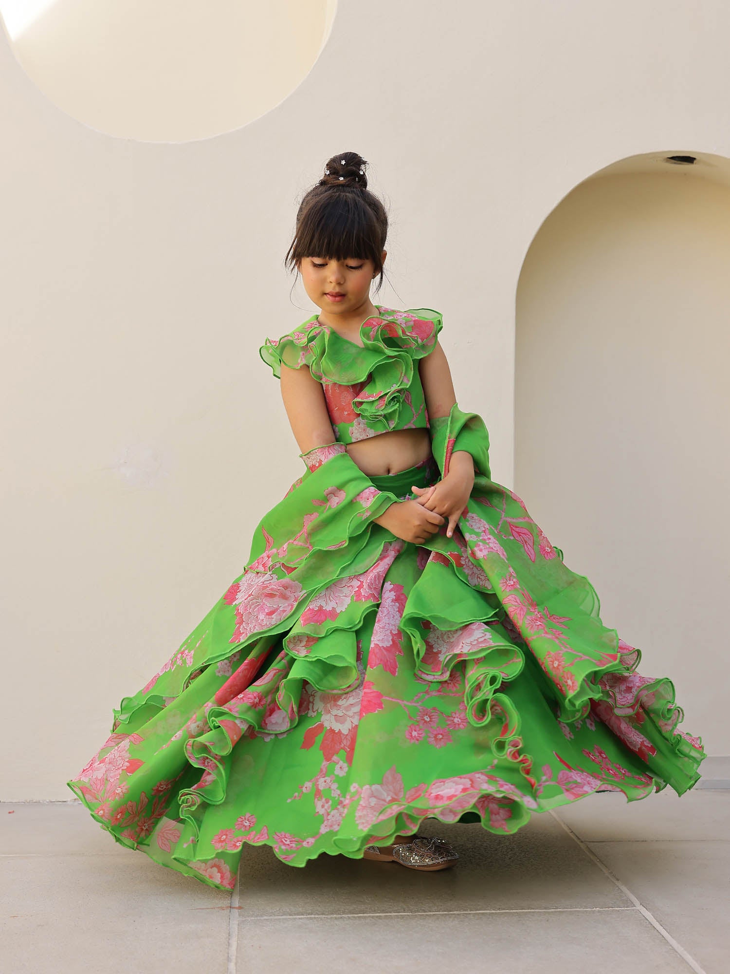 Little girl smiling in a green floral ruffle lehenga set with pink prints and organza dupatta, styled with a neat bun hairstyle.
