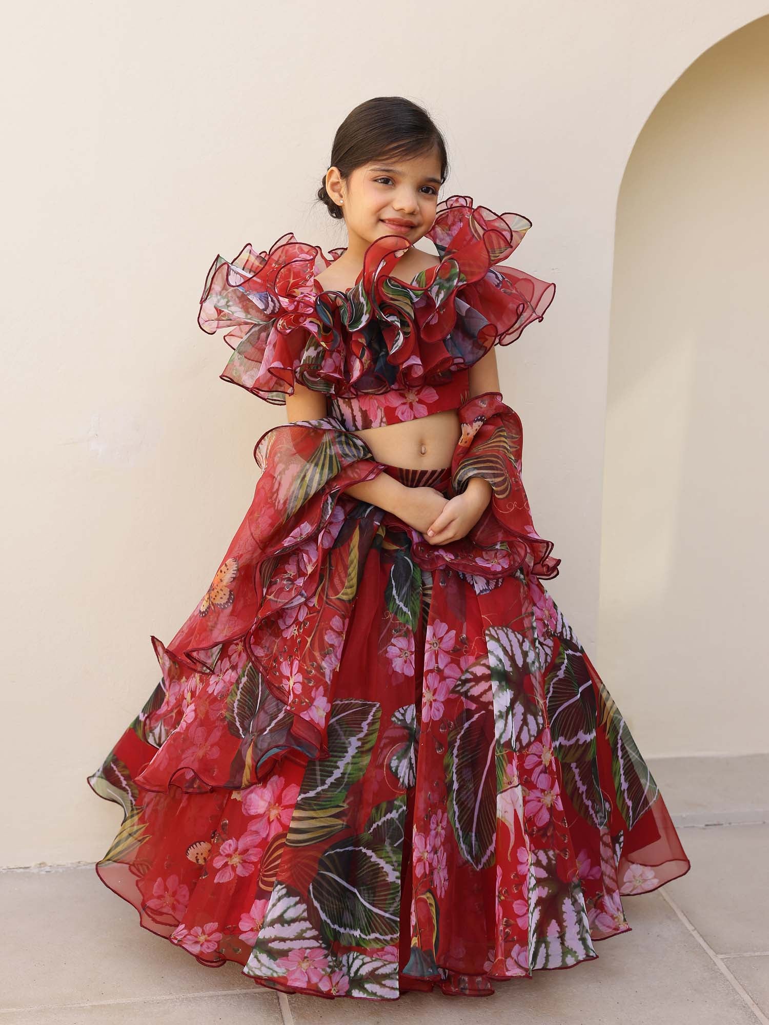 Little girl wearing a red-maroon floral organza lehenga with ruffled off-shoulder blouse.