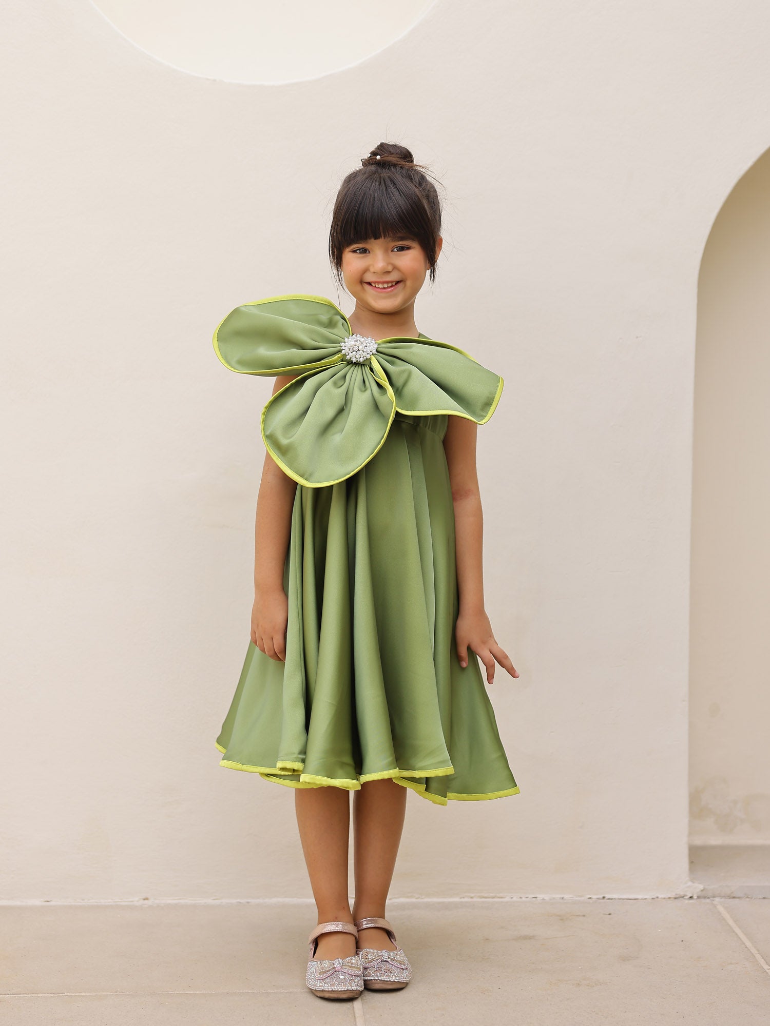 Young girl wearing a green dress with a large bow in front of a white wall.