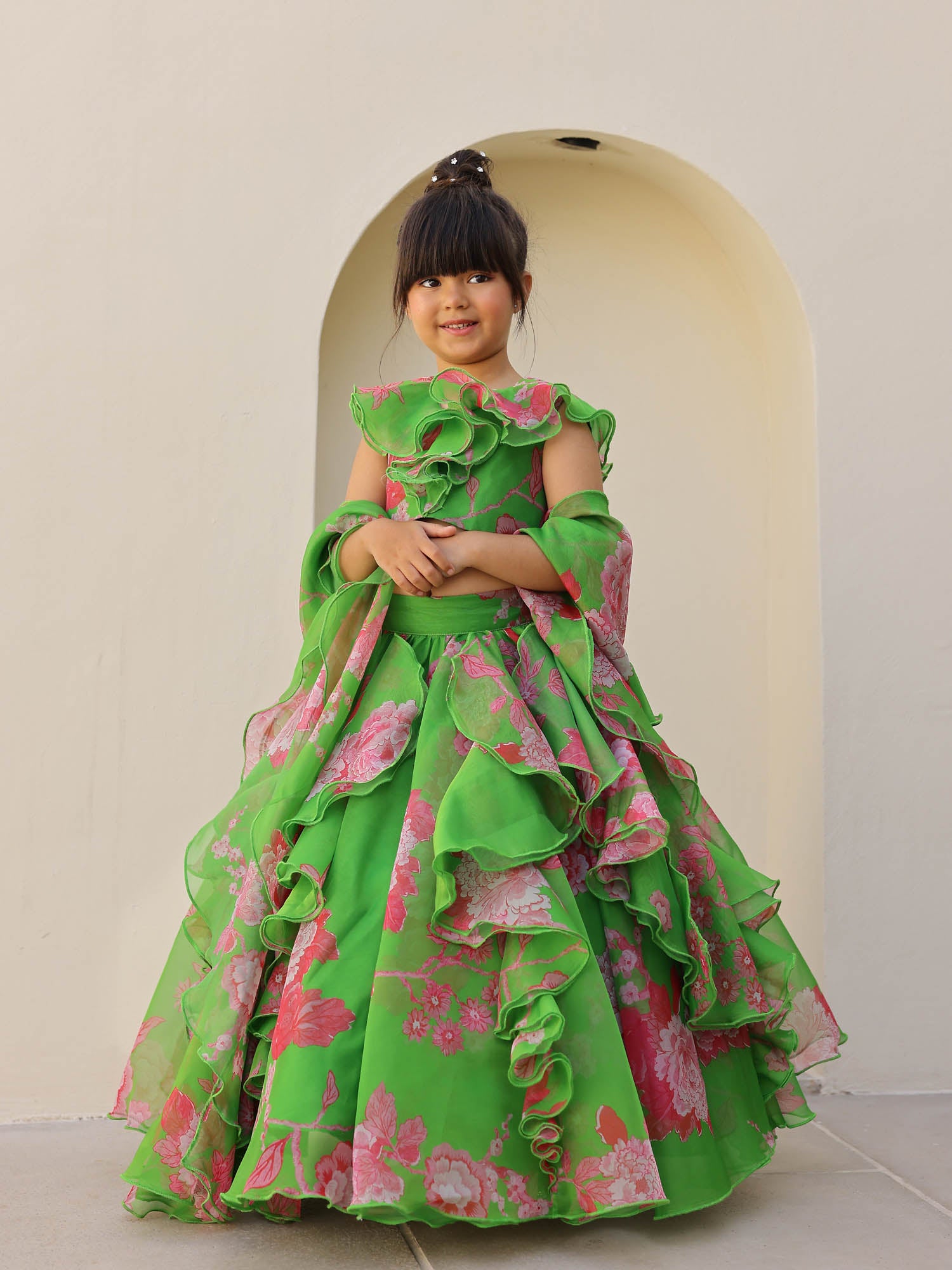 Little girl wearing a green floral organza lehenga with pink flower prints and layered ruffles, posing gracefully against a cream background.