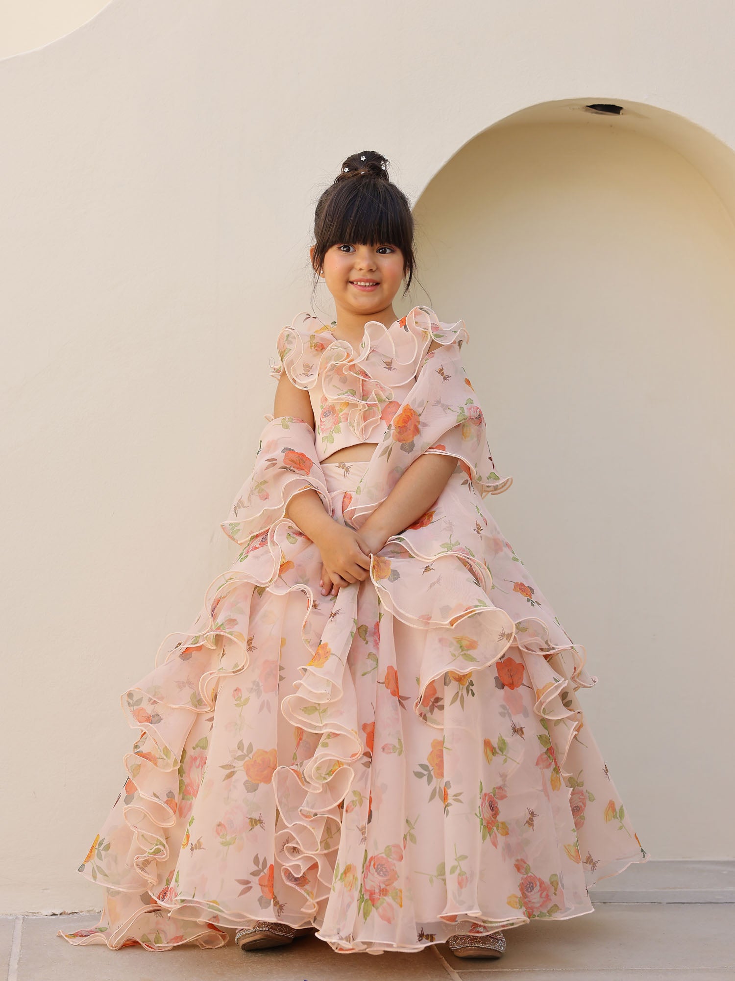 Little girl wearing a beautiful peach floral organza lehenga with orange flower prints and layered ruffles.